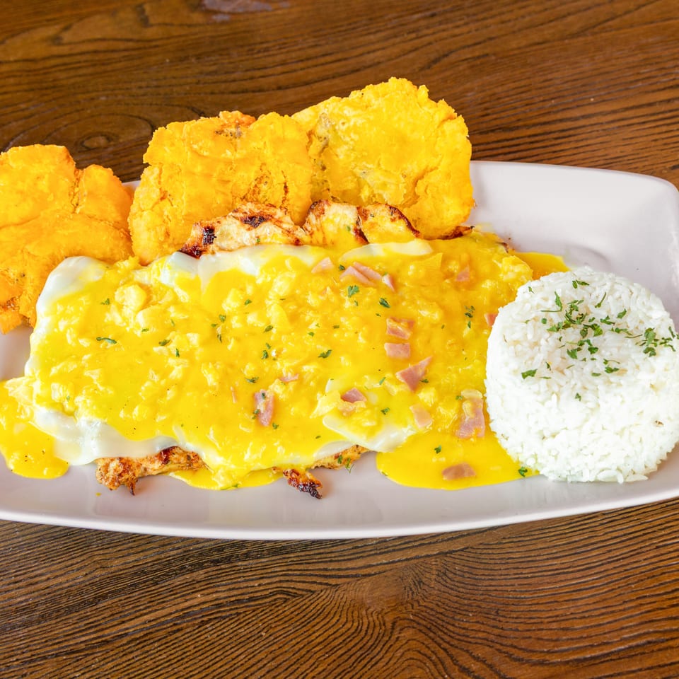 Grilled chicken topped with creamy yellow sauce, ham, white rice, and crispy tostones.