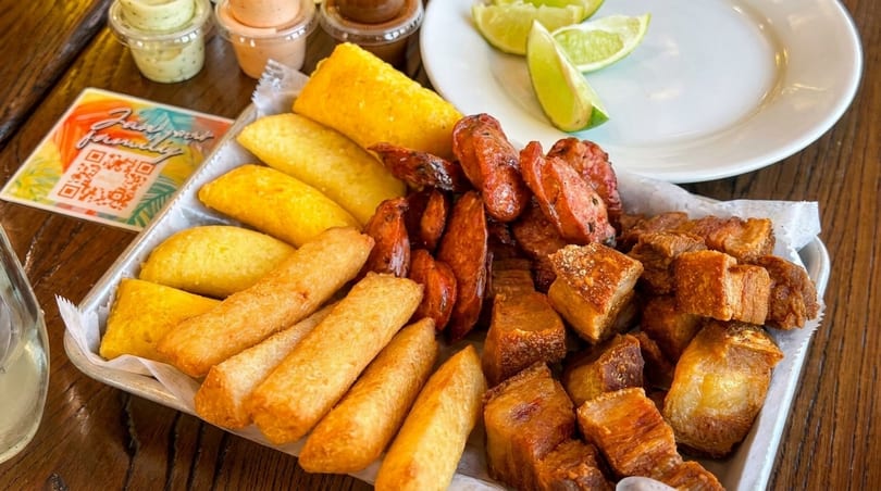 A tray of fried pork belly, sausages, empanadas, and yuca fries with dipping sauces and limes.