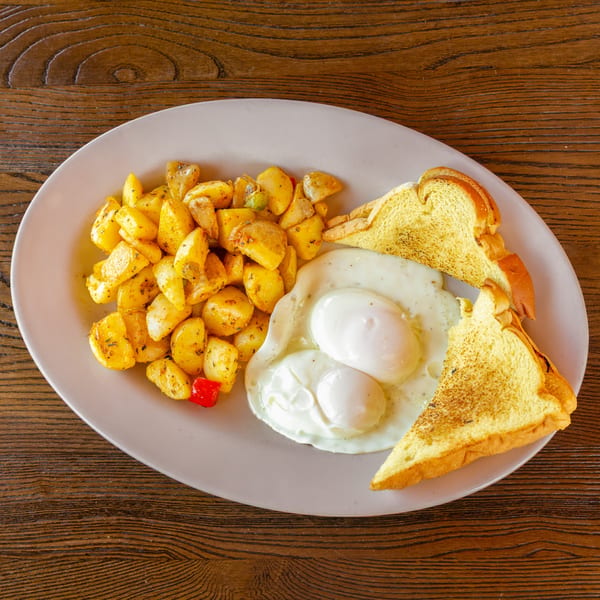 Breakfast plate with two fried eggs, seasoned home fries, and two slices of toast on a wooden table.