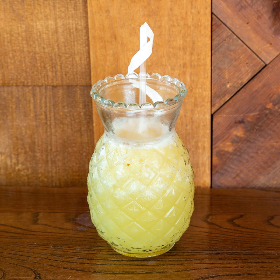 Pineapple-shaped glass filled with a yellow drink and a white straw on a wooden surface.