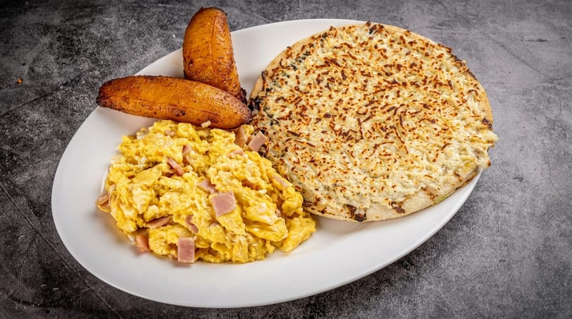 A white plate with scrambled eggs and ham, two fried plantains, and a cheesy arepa.