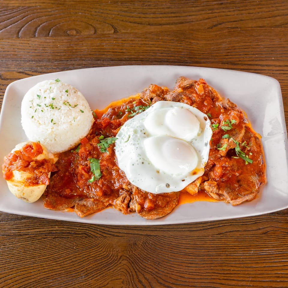 Steak with red sauce, two fried eggs, white rice, and a potato on a white plate.
