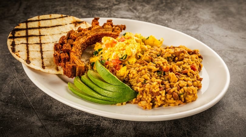 A white plate with a grilled arepa, chicharron, scrambled eggs, rice, and sliced avocado.