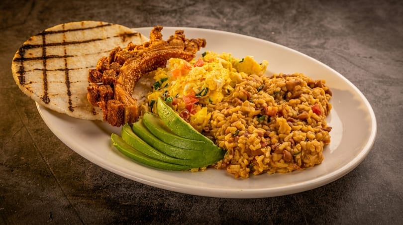 A white plate with a grilled arepa, fried pork belly, scrambled eggs, rice, and sliced avocado.