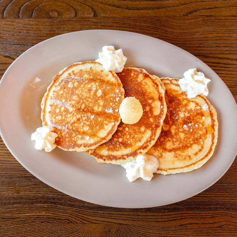 Three golden pancakes with syrup, powdered sugar, butter, and whipped cream on a plate.