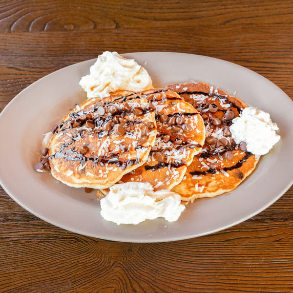 Pancakes topped with chocolate chips, coconut, chocolate drizzle, and whipped cream on a wooden table.